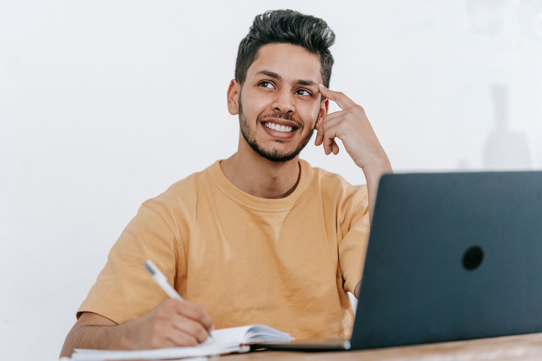 Jeune homme souriant, assis devant son ordinateur portable, qui prend des notes dans un carnet tout en réfléchissant à de nouvelles idées