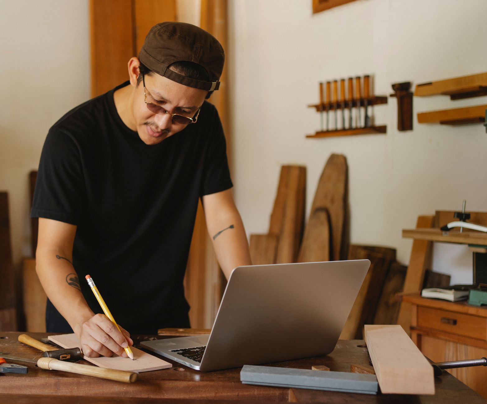Homme coiffé d’une casquette, penché sur un établi d’atelier, qui note quelque chose au crayon à côté d’un ordinateur portable