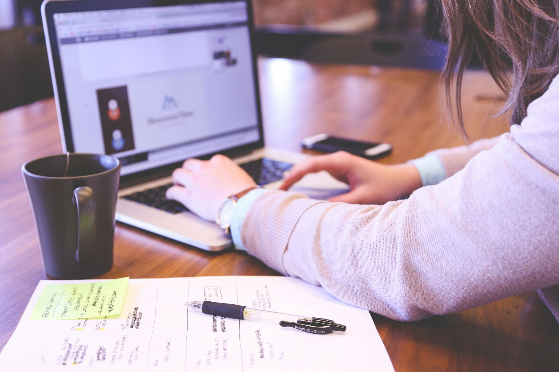 Femme qui tape sur un ordinateur portable, avec un carnet de notes, un stylo et une tasse posés sur un bureau en bois