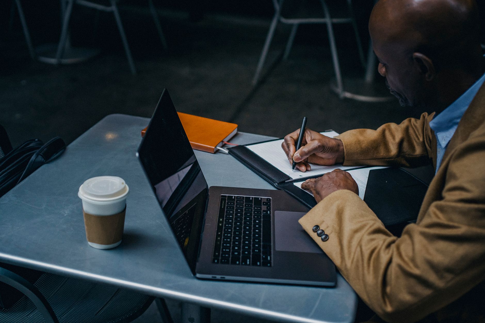Homme assis à une table, qui écrit des notes dans un carnet à côté d’un ordinateur portable et d’un gobelet de café