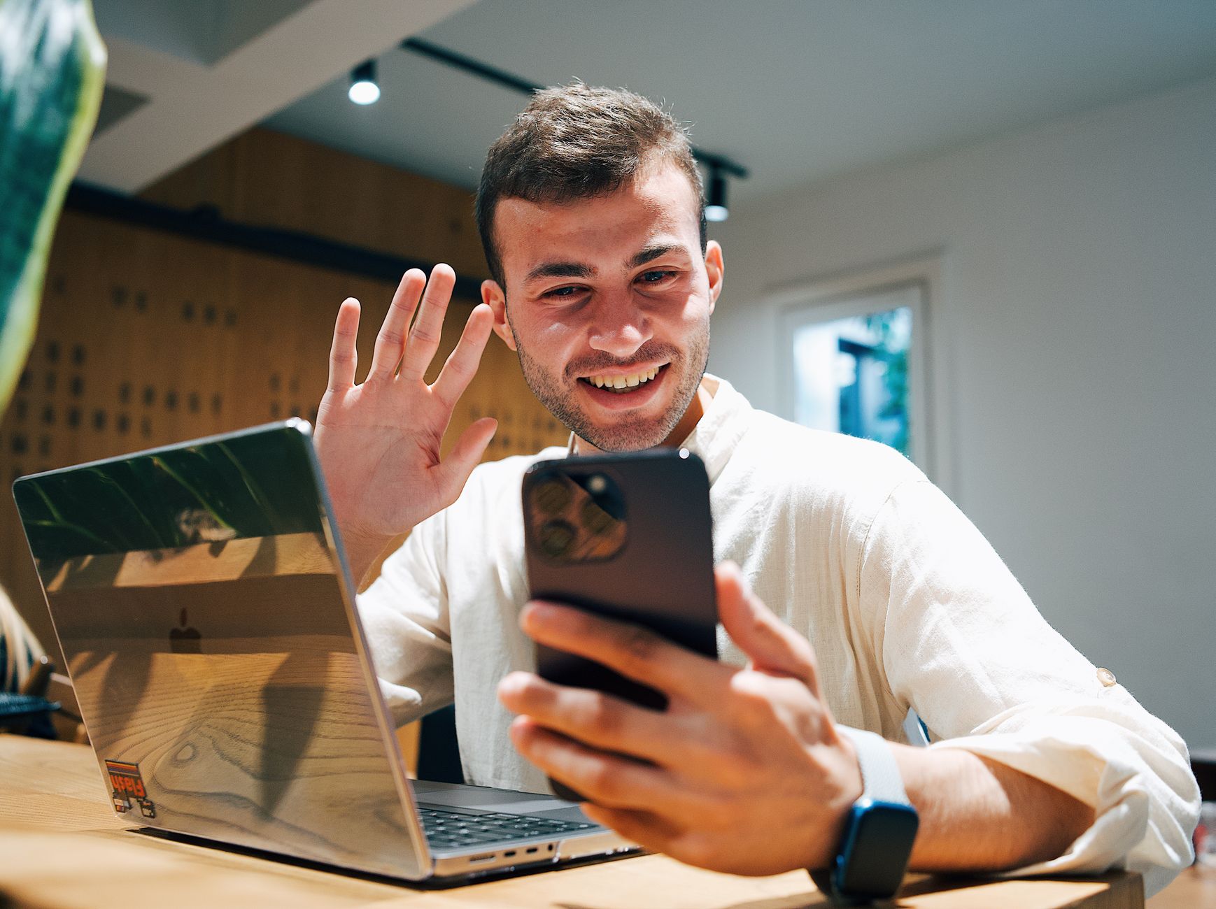 Un jeune homme souriant fait un appel vidéo sur son smartphone, assis devant un ordinateur portable, dans un bureau lumineux.