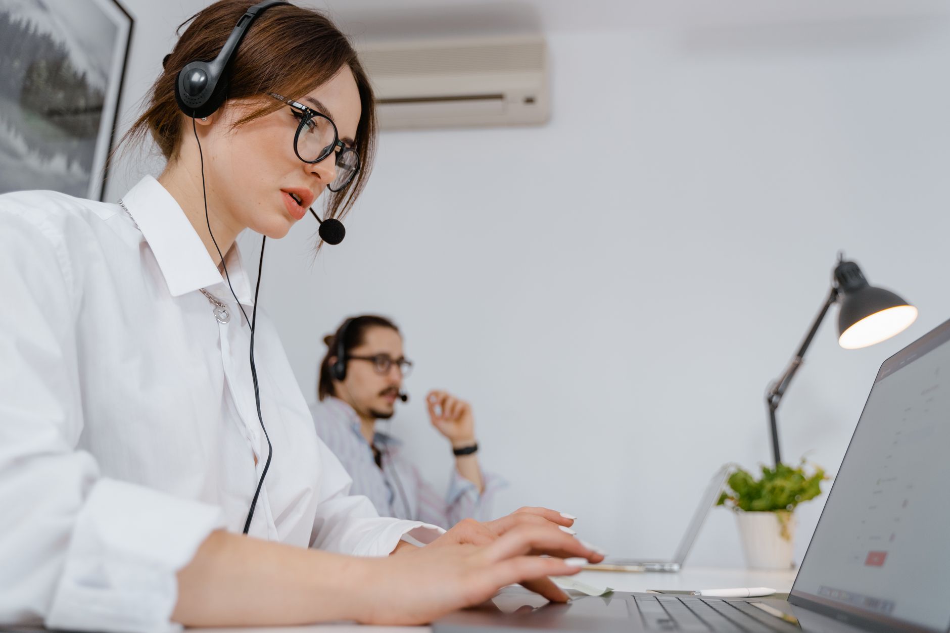 Deux personnes équipées de casques-micros travaillent sur ordinateur portable dans un bureau clair.
